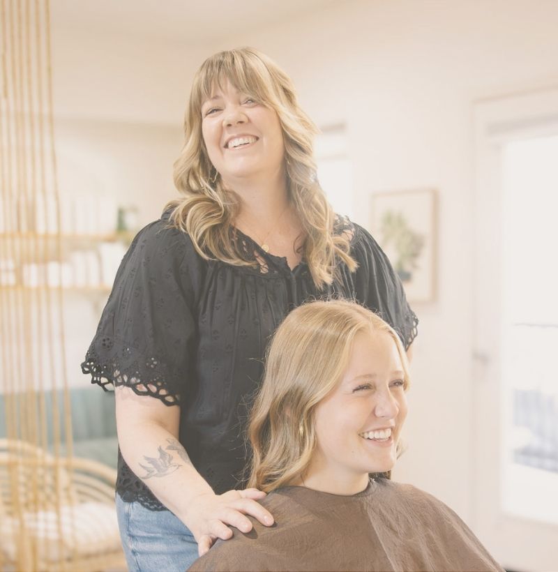 new guest experience 1 a woman sits in a hair styling chair getting her hair cut and styled while the stylist stands behind her smiling